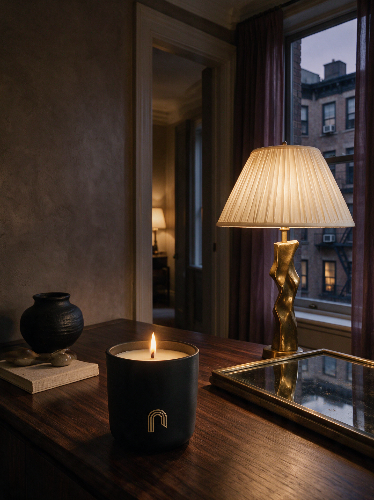 A lit matte black candle with a small Shelton Jaymes arch logo sits on a dark wooden table in a dimly lit room. A lit table lamp with a pleated cream shade and a gold sculptural base stands beside it, along with a mirror, a small black vase on a book, and a window looking out onto city buildings.
